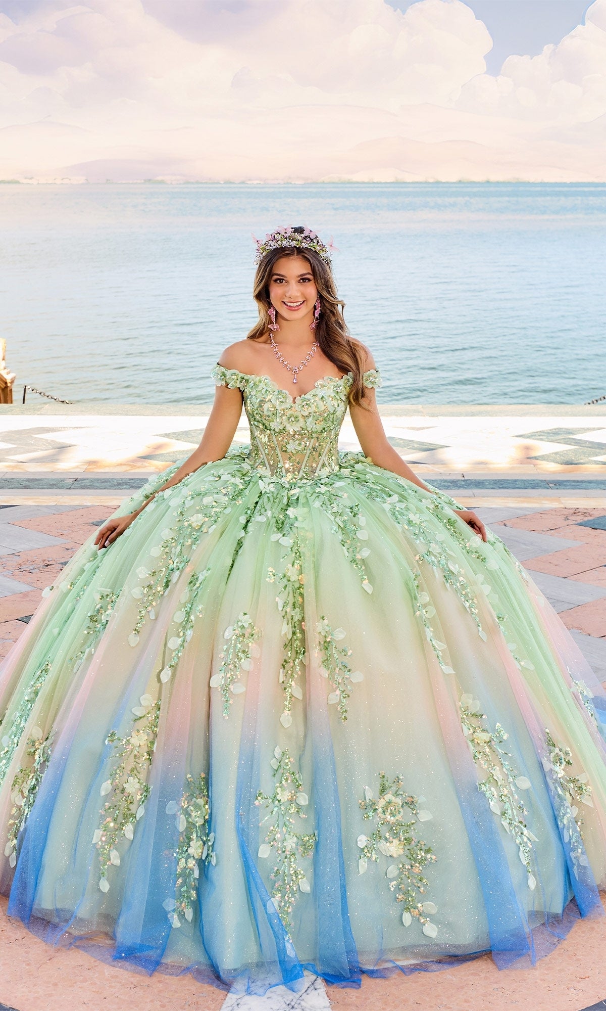 A young woman in the Quinceanera Dress PR30162 By Princesa, featuring an ombre design, floral embellishments, and an off-the-shoulder neckline, poses by a waterfront under a sunny sky.